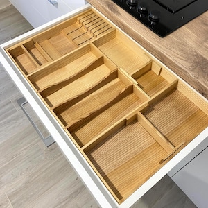 Könnte beinhalten: Offene weiße Küchenschublade mit einem individuellen Holz-Organizer. Der Organizer hat mehrere Fächer und Schlitze für Besteck, Utensilien und andere Küchenutensilien. Das Holz hat eine natürliche, hellbraune Farbe.