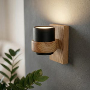 May include: A modern wall sconce featuring a black cylindrical light fixture mounted on a rectangular wooden base. The light is surrounded by a wooden ring and emits a warm glow. The sconce is attached to a gray wall.