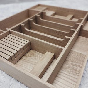 May include: A light oak cutlery tray with several compartments. The rectangular tray has slots for knives and other utensils. The wood grain is visible, and the design is simple and functional, suitable for a kitchen drawer.
