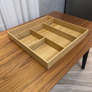 Può includere: Un organizer in legno color chiaro con più scomparti. L'organizer è rettangolare con diversi divisori che creano varie sezioni. È appoggiato su un tavolo di legno marrone. L'organizer è progettato per la conservazione e l'organizzazione.