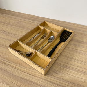 Puede incluir: Un organizador de cubiertos de madera con múltiples compartimentos, que contiene cubiertos y una espátula negra. El organizador es de madera clara y está sobre una superficie de madera.
