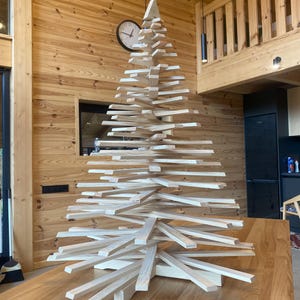 Peut inclure: Une sculpture décorative en forme de sapin de Noël composée de lattes de bois clair empilées horizontalement. Les lattes sont disposées en spirale et en dégradé pour former une silhouette conique, reposant sur une base en bois croisé.