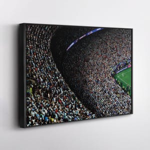 May include: A framed wall art print featuring a high-angle view of a crowded stadium during a soccer match. The stands are packed with spectators wearing light blue and white, with a green field visible in the corner and digital signage.