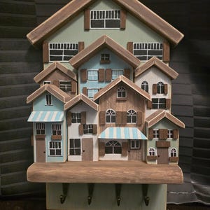 Op de afbeelding: Een houten sleutelhanger in de vorm van een rij huizen met een plank en haken. De huizen zijn geschilderd in tinten bruin, blauw en groen met witte accenten.