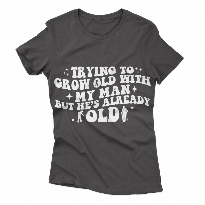 Puede incluir: Camiseta gris oscuro con texto blanco que dice "Trying to grow old with my man but he's already old". El texto est&aacute; en un estilo de fuente retro con estrellas y figuras de palitos de personas caminando con bastones.