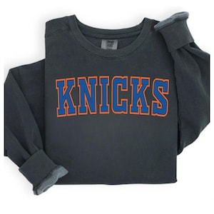 May include: Dark grey sweatshirt with the word "KNICKS" in blue block letters with orange trim. The long-sleeved shirt is folded, showing the cuffs and neckline. The shirt is made of a soft material.