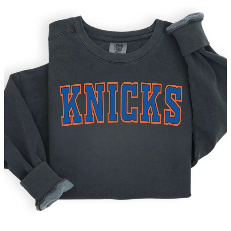 May include: Dark grey sweatshirt with the word "KNICKS" in blue block letters with orange trim. The long-sleeved shirt is folded, showing the cuffs and neckline. The shirt is made of a soft material.