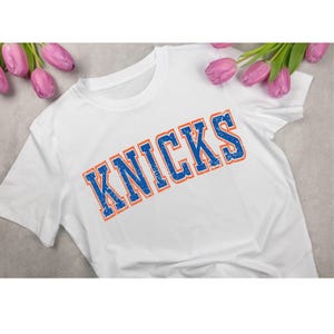 May include: White t-shirt with the word "KNICKS" printed in blue with an orange outline. The text has a distressed, vintage look. The shirt is displayed on a gray surface with pink tulips.