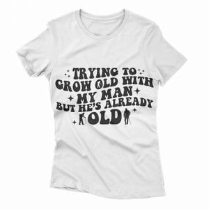 Puede incluir: Camiseta blanca con texto negro que dice "Intentando envejecer con mi hombre pero ya est&aacute; viejo". El texto est&aacute; en un estilo de fuente retro con estrellas y figuras de palitos de hombres mayores con bastones.
