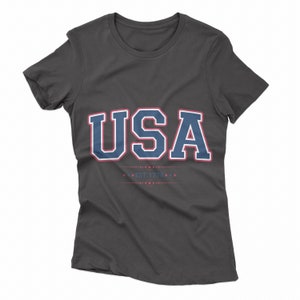 May include: A charcoal grey t-shirt with a navy blue and red "USA" graphic and the text "EST. 1776" in red.
