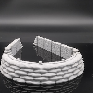 Op de afbeelding: Een wit, 3D-geprint model van een zandzakversterking met een houten plankenwandsectie. De zandzakken zijn in een halve cirkel gestapeld, waardoor een verdedigingsbarrière ontstaat.