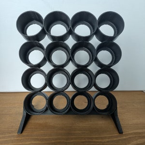 Puede incluir: Organizador de plástico negro con 16 agujeros redondos, diseñado para guardar objetos pequeños como bolígrafos, lápices o pinceles. El organizador tiene una base con dos patas.