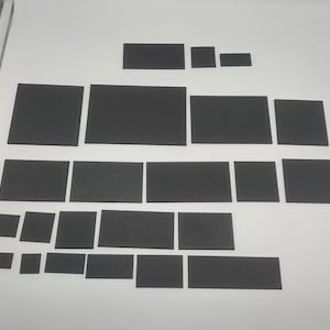 May include: A collection of black, rectangular foam pads of different sizes are arranged on a white surface. The pads are in various shapes, including squares and rectangles, and are evenly spaced.