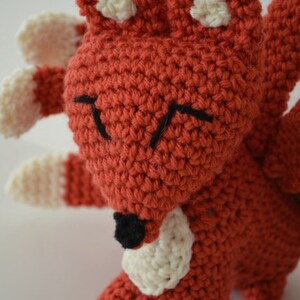 May include: A crocheted red fox with white accents and black eyes. The fox has multiple tails and is standing on a white surface.