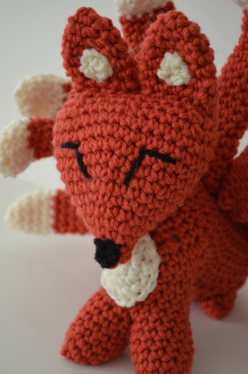 May include: A crocheted red fox with white accents and black eyes. The fox has multiple tails and is standing on a white surface.