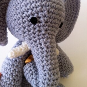 May include: A gray crocheted elephant stuffed animal with white tusks and black eyes. The elephant is sitting on a white surface.