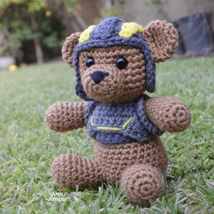 May include: A brown crocheted teddy bear wearing a dark gray helmet with yellow accents and a matching dark gray chest piece. The bear has black plastic eyes and a black stitched nose. The text Ami Amour is visible in the bottom left corner.