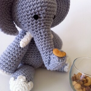 May include: A gray crocheted elephant toy holding a small orange snack in its trunk. The elephant has white crocheted accents on its ears, feet, and trunk. The elephant is sitting on a white surface.