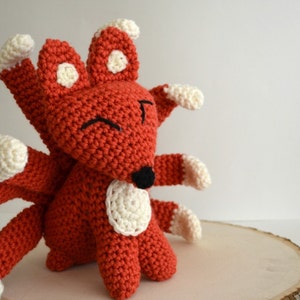 May include: A crocheted red fox with white accents and multiple tails. The fox has a black nose and smiling eyes.
