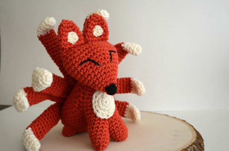 May include: A crocheted red fox with white accents and multiple tails. The fox has a black nose and smiling eyes.
