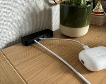 May include: A black cable organizer with four slots for cords, sitting on a wooden surface with white cords running out of the slots. A white wireless charging case is on the surface with a white cord running out of it. A green potted cactus is in the background.