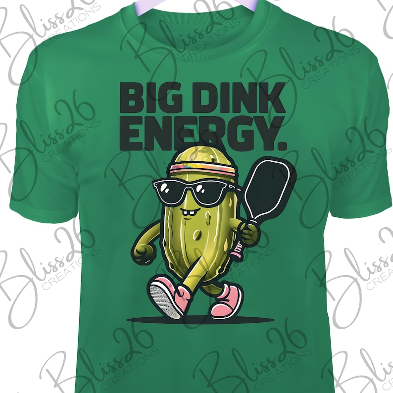 May include: Green t-shirt featuring a cartoon pickle wearing sunglasses and a headband, holding a pickleball paddle. The text "BIG DINK ENERGY." is printed on the shirt.