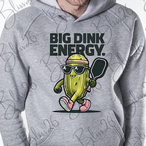 May include: A grey hoodie featuring a pickle character wearing sunglasses and a headband, holding a pickleball paddle. The text "BIG DINK ENERGY." is printed on the hoodie.