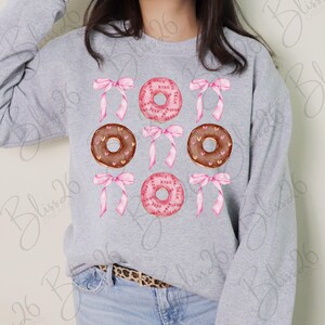 May include: A gray sweatshirt featuring a pattern of pink bows and donuts. The donuts are either pink with "xoxo" text or brown with heart sprinkles. The design is a Valentine's Day theme.