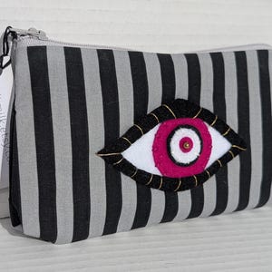 May include: A black and gray striped zippered pouch with an embroidered eye design. The eye features a white iris, a pink pupil, and gold-colored detailing. The tag reads: spiltmilk.etsy.com.