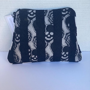 May include: Black and white zippered pouch with a skull lace design. The pouch features vertical black lace stripes alternating with white fabric, creating a striking contrast. The skull pattern is visible within the lace. The zipper is black.