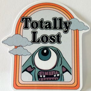 May include: A sticker with a retro design featuring the text "Totally Lost" above a cartoon cyclops monster. The design includes a rainbow arch, clouds, and a border with orange and brown tones.