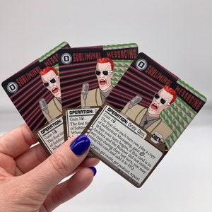 May include: Three illustrated playing cards with a red and black background. The cards feature a man with red hair and sunglasses wearing a beige shirt and holding a microphone. The text on the cards reads "Subliminal Messaging" and "Operation: Gray Ops".