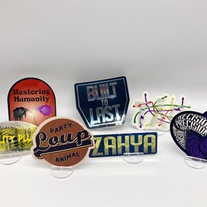 Könnte beinhalten: Eine Auswahl an bunten Aufklebern mit verschiedenen Designs und Texten, darunter "Restoring Humanity", "Built to Last", "Party Animal Loup", "Zahya" und "Precision".