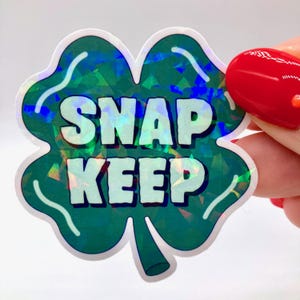 May include: A holographic sticker in the shape of a four-leaf clover. The sticker has a green background with iridescent accents and the words "SNAP KEEP" in white block letters. The sticker is held by a person with red nail polish.