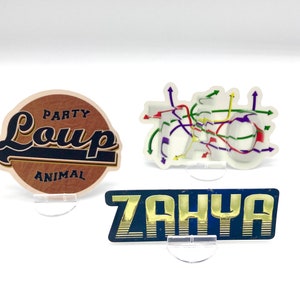 Könnte beinhalten: Drei dekorative Aufkleber mit klaren Acrylständern. Der erste Aufkleber ist braun mit einer braunen Ledertextur und trägt den Text "Party Animal Loup" in Schwarz und Weiß. Der zweite Aufkleber ist weiß mit einem bunten Design aus Pfeilen und Linien. Der dritte Aufkleber ist blau mit goldfarbenem Text, der "Zahya" lautet.