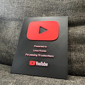 May include: A black plaque with a red YouTube play button logo. The text reads "Presented to Linus Knives For passing 70 subscribers" and the YouTube logo is at the bottom.