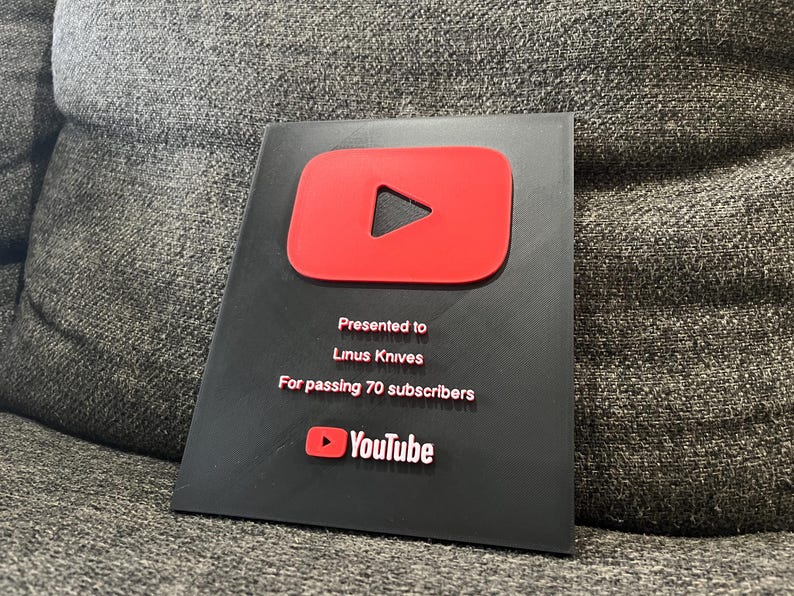 May include: A black plaque with a red YouTube play button logo. The text reads "Presented to Linus Knives For passing 70 subscribers" and the YouTube logo is at the bottom.