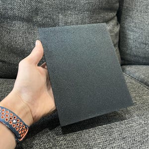 May include: A rectangular, black, textured panel is held in a hand. The panel has a matte finish and appears to be made of a solid material. The background is a gray upholstered surface.