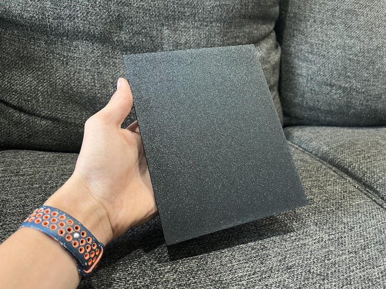 May include: A rectangular, black, textured panel is held in a hand. The panel has a matte finish and appears to be made of a solid material. The background is a gray upholstered surface.