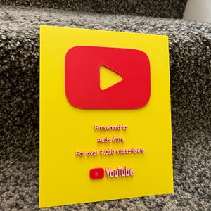 May include: A yellow plaque with a red YouTube play button logo. The plaque is presented to Auds Aura for over 2,000 subscribers. The YouTube logo is at the bottom. The plaque is on a textured surface.