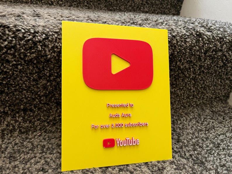 May include: A yellow plaque with a red YouTube play button logo. The plaque is presented to Auds Aura for over 2,000 subscribers. The YouTube logo is at the bottom. The plaque is on a textured surface.