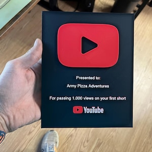 May include: A black plaque with a red YouTube play button logo. The text reads "Presented to: Army Pizza Adventures. For passing 1,000 views on your first short." The YouTube logo is at the bottom.