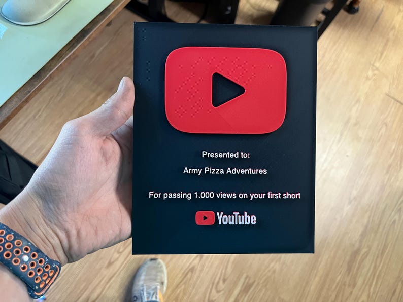May include: A black plaque with a red YouTube play button logo. The text reads "Presented to: Army Pizza Adventures. For passing 1,000 views on your first short." The YouTube logo is at the bottom.