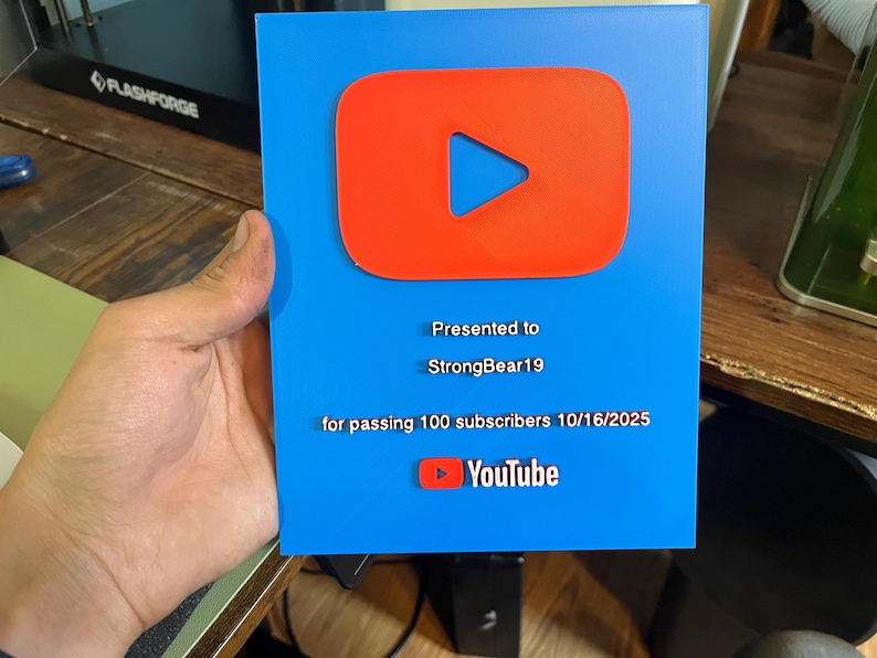 May include: A blue plaque with a red YouTube play button logo. The plaque reads "Presented to StrongBear19 for passing 100 subscribers 10/16/2025" and includes the YouTube logo.