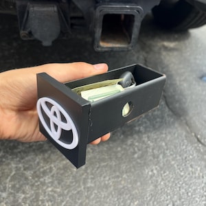 May include: Black metal storage box with a white Toyota logo, containing keys and cash. The box is held in front of a car's trailer hitch. The box has a rectangular shape with a circular hole on the side.