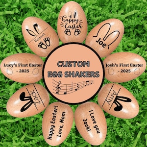 Pode incluir: Chocalhos de ovo de Páscoa de madeira com texto personalizado e desenhos de orelhas de coelho. Os ovos são dispostos sobre grama verde de Páscoa. O texto inclui "Feliz Páscoa", nomes e "Custom Egg Shakers".