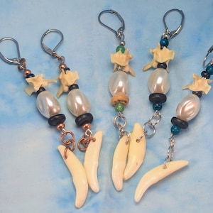 May include: Handmade earrings featuring bone, pearl, and bead accents. Each earring has a lever-back closure, a bone-shaped charm, and a combination of white pearls, black beads, and copper-colored accents. The earrings are displayed against a blue background.