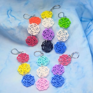 May include: A collection of colorful rose-shaped keychains. Each keychain features three stacked roses in various colors, including red, yellow, white, purple, black, blue, pink, and green, attached to a silver clasp.