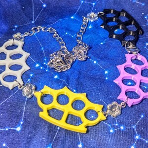 Puede incluir: Un collar multicolor con colgantes de nudilleras en blanco, amarillo, negro y rosa. Los colgantes están conectados por una cadena plateada y pequeños dijes de flores. El collar se exhibe sobre una tela azul con un patrón de constelaciones.