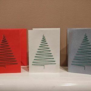 Puede incluir: Tres tarjetas de Navidad hechas a mano con un diseño de árbol de hilo verde. Las tarjetas son roja, blanca y gris.